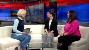 Three women sit on a white curved sofa in a TV studio, the host with a tablet interviewing two guests as large NYBERG screens loom behind them on red and blue panels.